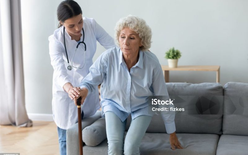 Young caretaker nurse doctor help injured disabled old grandma patient get up from sofa at homecare visit, female carer assist elderly woman hold walking cane stick, senior people healthcare concept